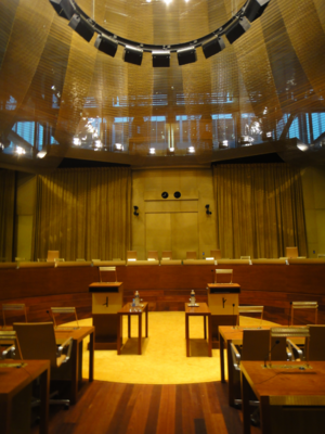 A courtroom in wooden ochery-brownish hues with a semi-circular table in the background with several armchairs behind it (the bench of judges), and two lecterns in front of them. Two rows of desks with chairs behind them are visible in the foreground.
